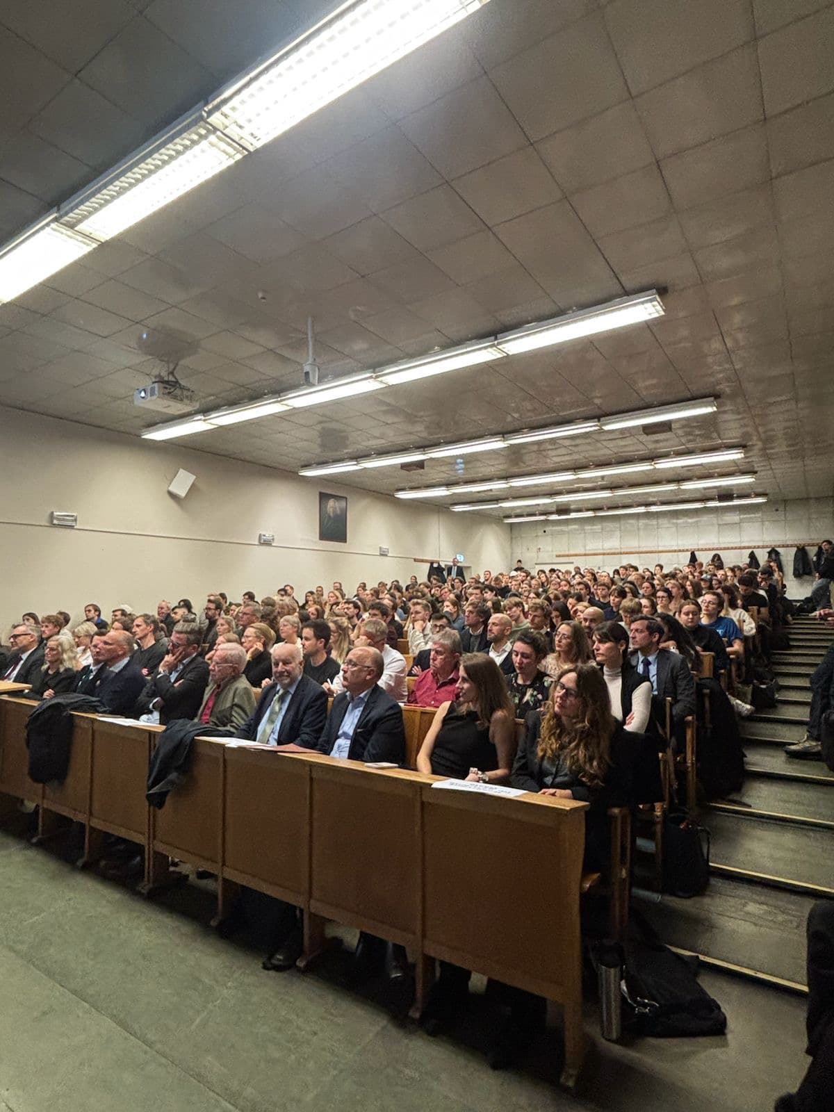 Großer, gut gefüllter Hörsaal während einer Veranstaltung. Reihenweise sitzen zahlreiche Menschen dicht nebeneinander und schauen nach vorn in Richtung Bühne oder Vortragende. Die Atmosphäre wirkt konzentriert; die Decke ist mit langen Neonlampen beleuchtet, und an der Seite sind Lautsprecher sowie ein Beamer angebracht.“